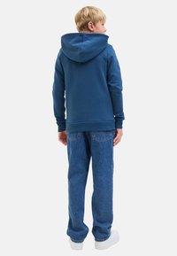 Sweat à capuche bleu avec cordon de serrage, texture douce et ourlet côtelé, associé à un jean bleu ample et des baskets blanches.