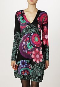 Nainen pukeutuneena mustaan pitkähiharaiseen mekkoon, jossa on värikkäitä abstrakteja ympyrä- ja kukkakuviota vaaleanpunaisissa, violeteissa, vihreissä ja sinisissä sävyissä.