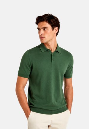 Hombre con camiseta polo de manga corta verde oscuro con botones y pantalones beige claros, de pie con las manos en los bolsillos mirando de lado.