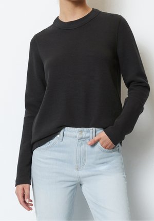 Person wearing a black textured long-sleeve top tucked into light blue high-waisted jeans, with one hand in pocket.