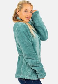 Giacca in pile color teal con colletto alto e maniche lunghe, caratterizzata da una texture morbida e un design aderente. Inclusi taschini laterali.