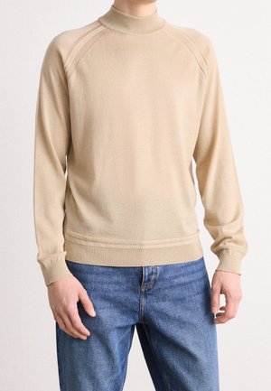 Mann trägt einen beigefarbenen Pullover mit langen Ärmeln, geripptem Kragen und Manschetten, kombiniert mit blauen Jeans, steht vor einem schlichten hellen Hintergrund.