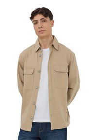 Camicia button-up beige con due tasche sul petto, colletto a punta e maniche lunghe, indossata sopra una maglietta bianca. I jeans blu completano l'outfit.