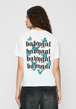Une femme aux cheveux bouclés porte un T-shirt blanc avec le texte "babygal" répété en noir et des papillons bleus imprimés au dos.