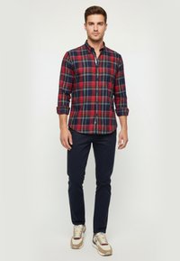 Camisa de cuadros roja y marina, de material de algodón, con mangas largas arremangadas, combinada con pantalones oscuros y zapatillas con acentos beige.