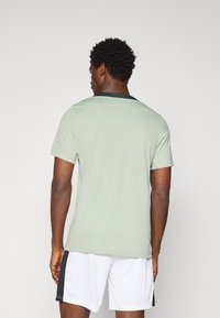 T-shirt de sport vert clair à manches courtes et encolure noire, fabriqué dans un tissu respirant. Porté avec un short blanc avec une rayure noire.