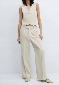 Veste beige à boutons avec un design raccourci, associée à un pantalon beige taille haute à jambes larges et des chaussures beiges à bout pointu.