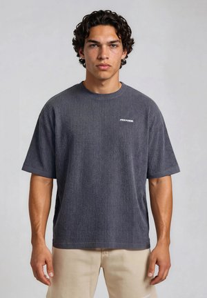 Ung mand med krøllet hår iført en ribstrikket mørkegrå kortærmet skjorte og lyse beige bukser, stående mod en ensfarvet grå baggrund.