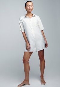 Kratka rokavi, svetlo bež srajčna obleka s prsno žepom, narejena iz mehkega blaga, z sproščenim krojem in minimalističnim dizajnom.