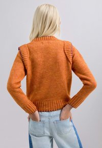 Suéter de punto naranja con detalles acanalados en el cuello y los puños, combinado con jeans de color azul claro, mostrando un ajuste casual.