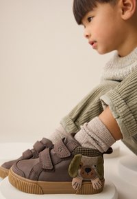 Braune Klettverschluss-Sneaker mit einem Hundegesicht-Design an der Seite, gefertigt aus glattem Material mit strukturiertem Akzent. Gummisohle und gepolsterter Kragen.