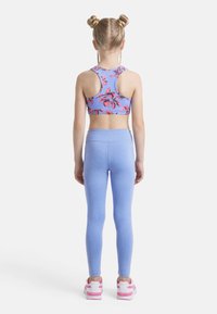 Νεαρή κοπέλα που στέκεται γυρισμένη με την πλάτη, φορώντας μπλε αθλητικό τοπ με λουλούδια, ταιριαστό μπλε κολάν, λευκά αθλητικά παπούτσια με ροζ λεπτομέρειες και χτενισμένο με δύο κότσους.