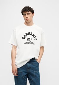 Νέος άνδρας που φοράει λευκό t-shirt Carhartt WIP με μαύρο κείμενο, συνδυασμένο με μπλε τζιν, στέκεται μπροστά σε ένα απλό φόντο.