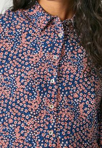 Blommig skjorta med knappar som har en marinblå bakgrund med röda och vita blommönster, lätt tygstruktur och vita knappar.