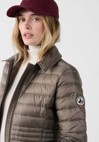 Gevoerde bruine jas met een kraag en rits, met een logo-opdruk op de mouw. Model draagt een bordeauxrode pet en een witte coltrui.