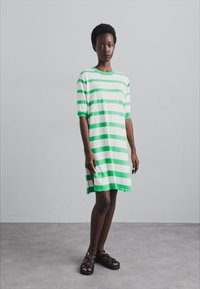 Abito a maniche corte a righe in bianco e verde, con una vestibilità ampia e scollatura rotonda. Indossato con sandali neri, su uno sfondo neutro.