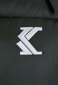Juodas audinys su lygiu paviršiumi, turintis baltą siuvinėtą logotipą su geometriniais raštais, išdėstytu ryškiai centre.
