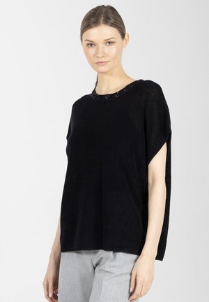 Kobieta o jasnej karnacji i brązowych włosach nosi luźny czarny krótki sweter z dzianiny i jasnoszare spodnie, stojąc na tle jednolitego jasnego tła.