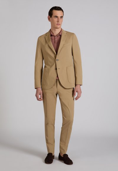 Homme portant un costume beige avec des mocassins marron et un polo mauve, debout devant un fond uni gris clair.