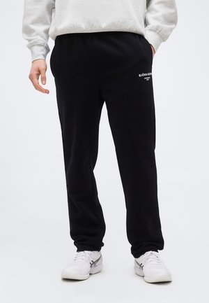 Mees, kes kannab musti Björn Borg dressipükse, valgeid tossusid ja heledat halli dressipluusi, seisab ühevärvilise tausta ees.