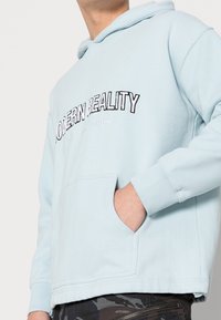 Světle modrá mikina s kapucí a přední kapsou ve tvaru klokana, vyšívaným nápisem „MODERNÍ REALITA“ a žebrovanými rukávy. Vyrobená z měkké bavlněné směsi.
