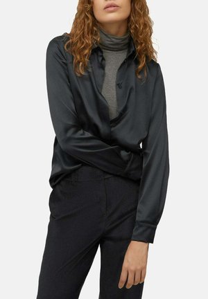 Femme portant une chemise noire en satin boutonnée sur un col roulé gris, associée à un pantalon noir, debout devant un fond uni.
