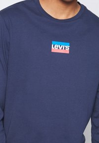 Mies, joka käyttää tummansinistä pitkähihasta paitaa, jossa on pieni Levi's-logo valkoisena, sinisenä ja punaisena rinnan keskellä.
