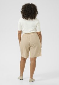 Shorts beiges avec une taille élastique, fabriqués en tissu léger, portés avec un haut blanc ajusté et des chaussures beiges à enfiler.