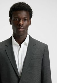 Grau strukturiertes Blazer über einem weißen Hemd mit spitzem Kragen. Das Blazer verfügt über eine einzelne Brusttasche und eine taillierte Passform.