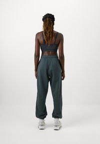 Sötét szürke sportmelltartó széles pántokkal, laza, keskeny szabású sötétzöld tréningnadrággal, fehér sportcipővel és bokazoknival viselve.