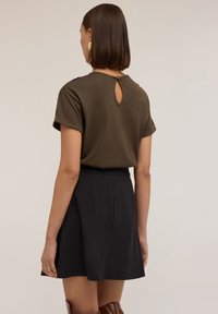Olivově zelený top s krátkým rukávem a detaily ve tvaru klíčové dírky na zadní straně, spárovaný s černou plisovanou sukní. Hladká textura látky a volný střih.
