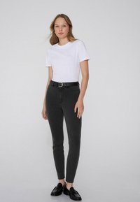 Jeans negros ajustados con cintura alta, con pasadores para cinturón, bolsillos frontales y una tela texturizada. Combinados con una camiseta blanca lisa y zapatos negros.