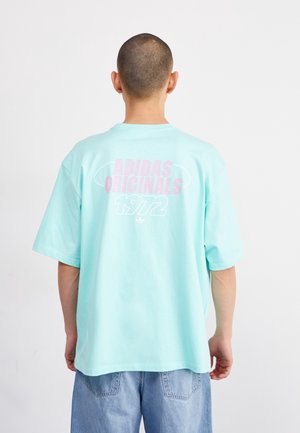 Person mit rasiertem Kopf, die ein hellblaues Adidas Originals 1972 T-Shirt und hellblaue Jeans trägt, von hinten vor weißem Hintergrund gezeigt.