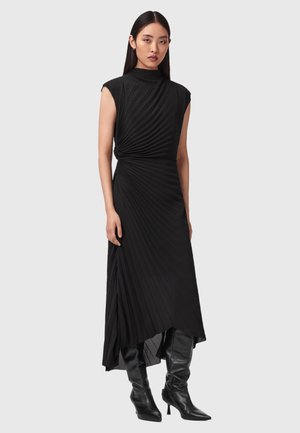 Kvinde iført en sort, ærmeløs midi-kjole med plisseret nederdel og høj hals, kombineret med sorte støvler med høje hæle, der går til knæene, stående foran en ensfarvet baggrund.