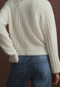 Pull blanc texturé avec des côtes verticales et des épaules tombantes, associé à un jeans en denim bleu comportant des poches arrière et une taille mi-haute.