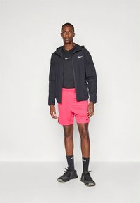 Nike'i spordirõivad: must lukuga jakk, must vormis särk, roosad lühikesed püksid, mustad sokid, mustad tossud. Lihtne, funktsionaalne disain.