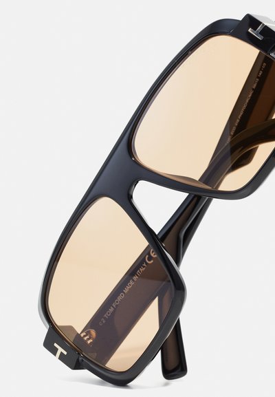 Gafas de sol negras cuadradas con lentes marrones claros. Cuentan con marcos gruesos y atrevidos, y un distintivo logo en forma de "T" en la parte frontal. Fabricadas en Italia.