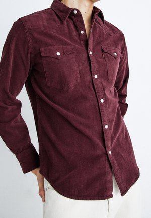 Homme portant une chemise en velours côtelé rouge foncé à manches longues, boutonnée, avec deux poches poitrine et des boutons-pression blancs.