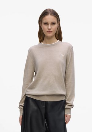 Kvinne iført beige genser med rund hals og lite hvitt logo på brystet og løse svarte skinnbukser, stående mot hvit bakgrunn.