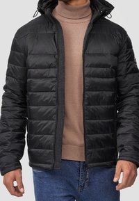 Veste matelassée noire avec capuche, dotée d'une fermeture éclair frontale, de poignets élastiques et d'un matériau léger, portée sur un pull col roulé beige clair.