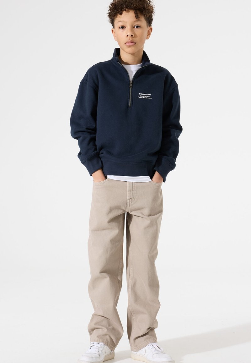 Sudadera azul marino con cremallera y cuello alto, combinada con pantalones rectos beige y zapatillas blancas. Presenta texto bordado en el pecho.