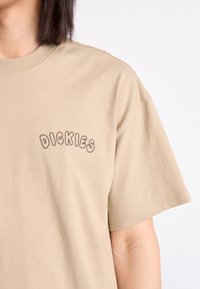 Smėlio spalvos medvilninė marškinėliai su trumpomis rankovėmis, su maža, išlenkta juoda "DICKIES" logotipu kairiajame krūtinės kampe. Lygus paviršius ir laisvas pjūvis.