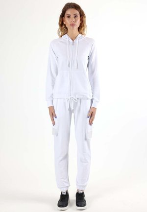 Une femme porte un sweat à capuche blanc zippé et un pantalon de jogging assorti avec des poches latérales, ainsi que des baskets noires, sur un fond blanc uni.