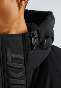 Svart pufferjacka med ett texturerat tyg och framträdande varumärkeslogga. Har en dragsko stängning och en hög krage för extra värme.