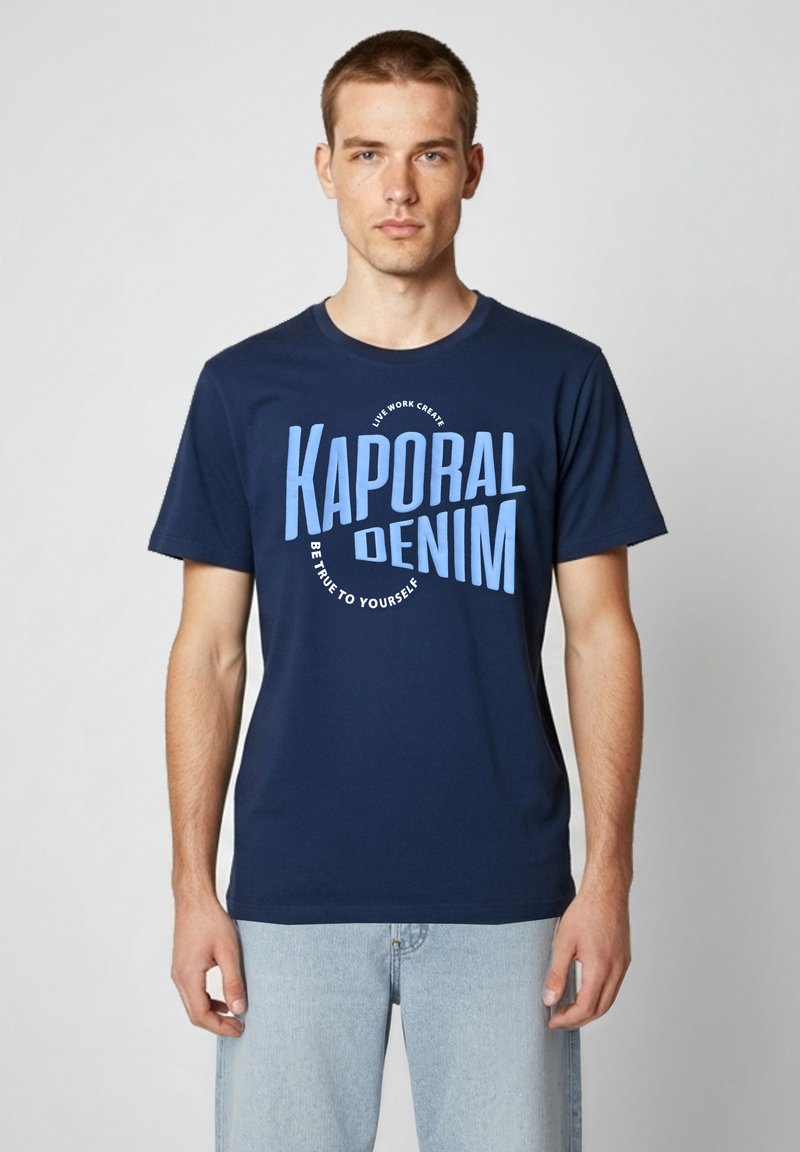 Jeune homme portant un t-shirt Kaporal Denim bleu marine avec texte bleu clair et jean bleu clair, debout devant un fond uni.