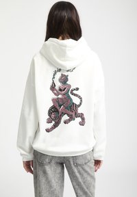 Hoodie blanc avec un design graphique audacieux représentant une figure chevauchant un tigre, agrémenté d'accents roses et turquoise. Fabriqué à partir d'un tissu doux et texturé.