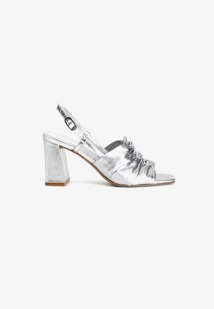 Sølvmetallic slingback sandal med blokhæl, med sammensnøring foran og firkantet tådesign. Detalje med spænde.