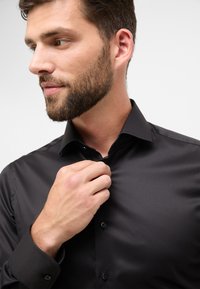 Camicia nera a maniche lunghe con colletto all'americana. Tessuto liscio, con un singolo bottone anteriore e bottoni a contrasto ai polsini.