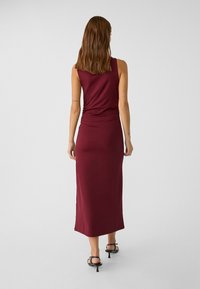 Bordeaux, mouwloze maxi-jurk met een aangesloten lijfje en gedrapeerde details bij de taille, gemaakt van zachte stof, gecombineerd met zwarte hoge hakken.