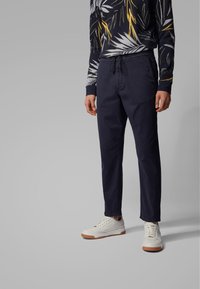 Navy drawstring pants with a smooth texture, paired with a patterned long-sleeve shirt in dark and bright colors, and white sneakers with gum soles.